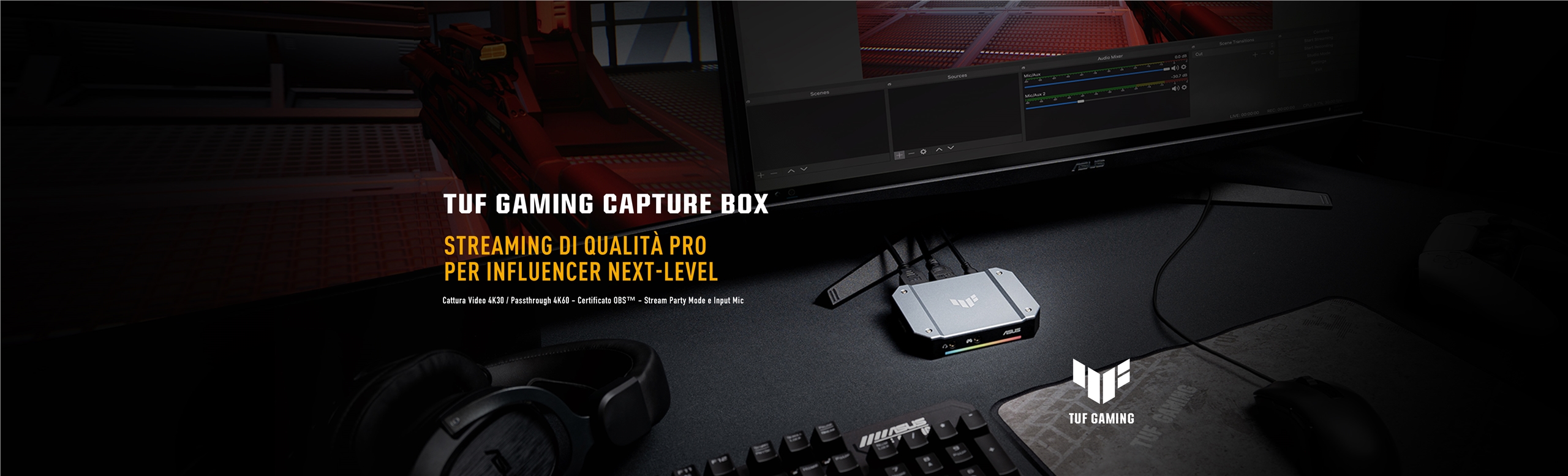 L'immagine mostra il box esterno per lo streaming ASUS TUF Gaming Capture Box con lo sfondo un moniotr di editing e delle periferiche gaming in primo piano